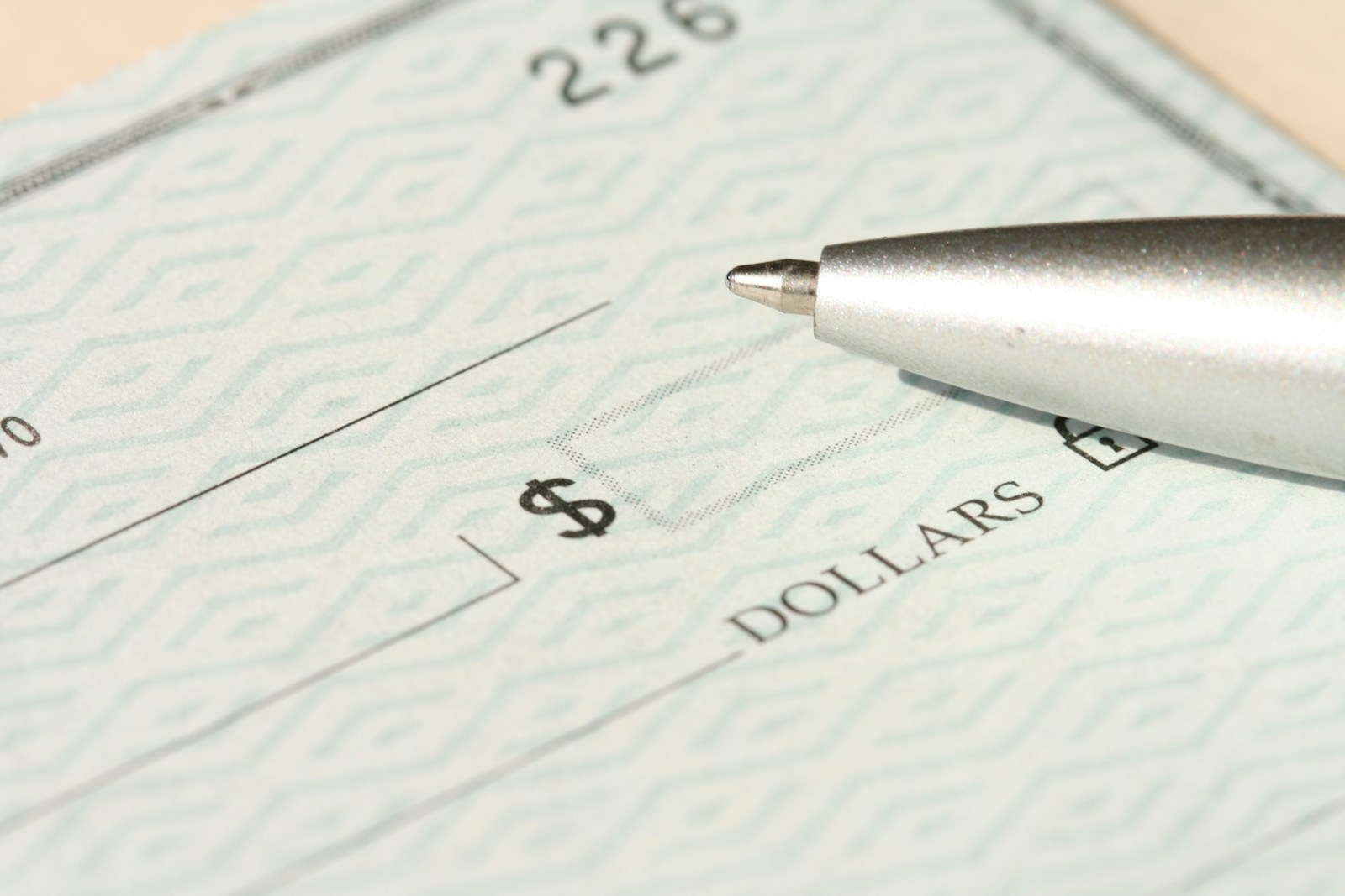 A close-up, high-resolution image of a blank check with a security pattern, showing the dollar sign box and part of the “DOLLARS” line. A metallic ballpoint pen rests near the writing area.